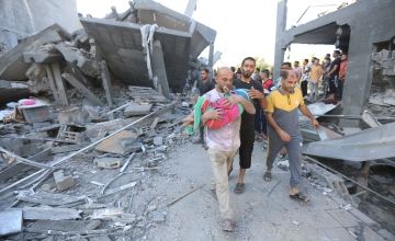 Man holding child walking through rubble and debris in Gaza followed by other people.