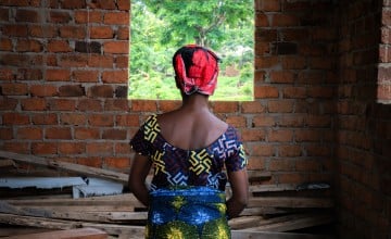 A woman in a colourful dress and headscarf looks out of a window in a brick building.