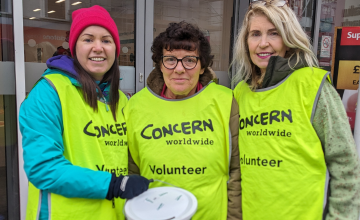 Three women stand together with a collection bucket wearing high-vis vests with a Concern logo and volunteer written on the front.
