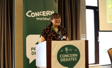 A person stands behind a podium with a microphone, delivering a speech. The podium displays a banner reading 'CONCERN DEBATES' with a microphone and speech bubble icon. Behind them is a 'CONCERN worldwide' banner and part of a projection screen.