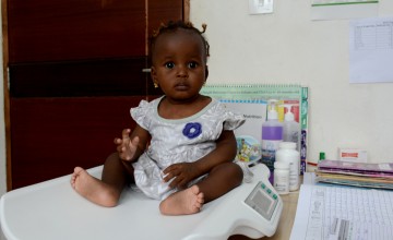 A young child in a light-colored dress sits on a digital weighing scale in a clinic setting. Behind her are nutrition charts, bottles, and paperwork on a desk, indicating a health check-up.