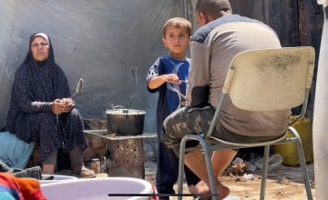 Three people in an outdoor setting: one sitting on the ground to the left holding a spoon, a child in the center interacting with a pot on a makeshift stove, and another person seated on a chair to the right facing away. Various items, including a plastic container, are scattered around them.