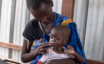 A woman gently feeds a young child sitting on her lap. The child appears weak and is dressed in a light pink shirt. They are seated on a bench in a shelter with metal panel walls. The woman wears a patterned blue shawl with yellow accents.