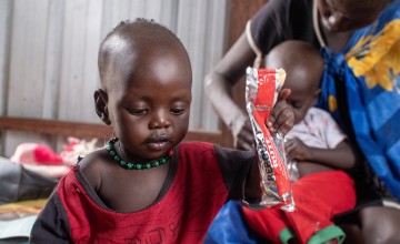 A young child wearing a red shirt and green beaded necklace holds a packet of therapeutic food while looking down. In the background, a woman feeds another child on her lap, sitting in a shelter with metal walls.