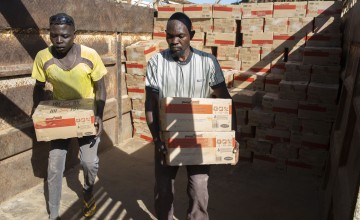 Two individuals carrying aid boxes in a storage area, surrounded by stacks of similar boxes. 
