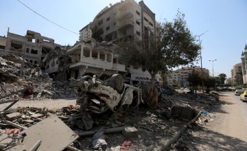 Scene of destruction with collapsed buildings and scattered debris. An overturned car is partially buried under rubble. Damaged structures and a lone tree are visible in the background under a clear daytime sky.