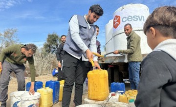 Several people filling yellow and white containers with water from a large white water tank labeled 'cesvi' in an outdoor setting under a clear blue sky