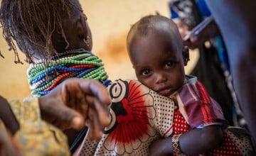 A young child looks directly at the camera while being carried on a woman’s back, wrapped securely in a brightly patterned cloth. The woman wears multiple colorful beaded necklaces and has braided hair, partially visible from behind.