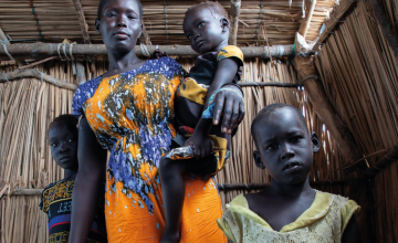 A woman stands inside a shelter made of sticks, holding a small child on her arm. Two other children stand close beside her, looking directly at the camera. The woman wears a brightly patterned orange and blue dress, and the children are dressed in colorful clothes.