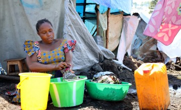 A woman sits in a displacement camp in a colourful dress surrounded by plastic containers.