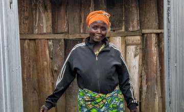 A woman in a colourful skirt and headscarf smiles into the camera in front of a wooden structure.