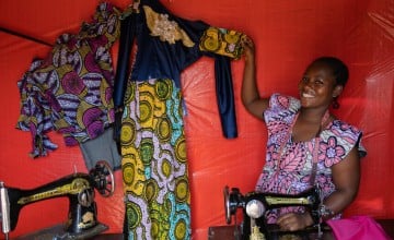 Woman smiling while seated at a sewing machine, holding up a colorful dress with yellow, blue, and green patterns and lace detailing. A second garment with purple and pink designs hangs nearby against a red fabric backdrop