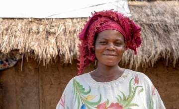 Person wearing a white garment with colorful floral patterns and a red patterned headwrap, standing in front of a traditional thatched-roof structure made of natural materials