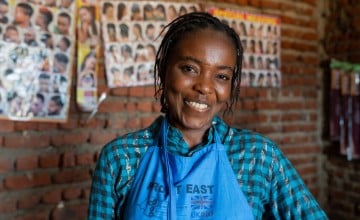 Smiling woman with braided hair wearing a blue apron with the UK aid logo, standing in front of a brick wall with hairstyle posters in the background