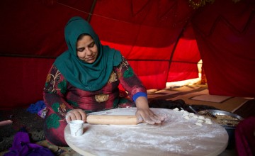A woman wearing a patterned dress and green headscarf rolls dough in an orange tent.