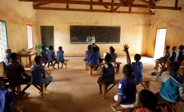 A light and airy classroom full of children being taught in front of a chalkboard. Some of the children are raising their hands.