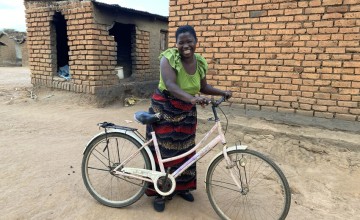 A smiling woman in ag reen top and red patterened skirt stands with a white bicycle  outside a brick building.