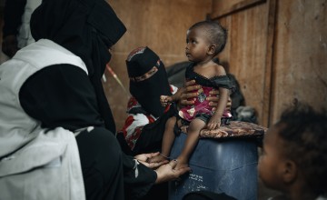 healthcare worker dressed in black clothing with a white vest is examining a small child seated on a blue plastic container. Another person wearing patterned clothing and a black face covering supports the child. The child is wearing a red and black outfit and appears thin, with visible ribs and limbs. The setting looks modest, with a brown wall in the background and another child partially visible in the foreground.