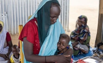 A mother in a red dress and blue scarf, sits on a chair, holding her baby at a health centre in Chad. Behind her other mothers wait holding their babies.