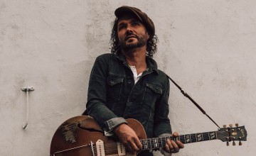 A man stands with a guitar against a white wall