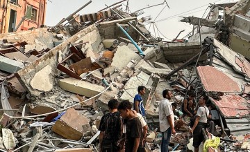 Several people stand and look through piles of rubble where nearby buildings have collapsed.