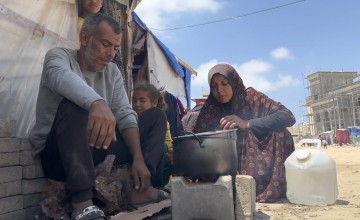A man, woman and child sit around a pot cooking on a makeshift fire on cinder blocks with a tent in the background.