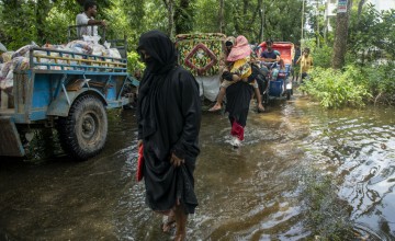 Flooded rural road with people wading through water near trucks and rickshaws after heavy rain.