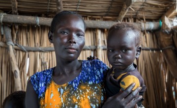 A woman holds a young child while standing inside a building made of wooden poles and woven reeds, both facing the camera.