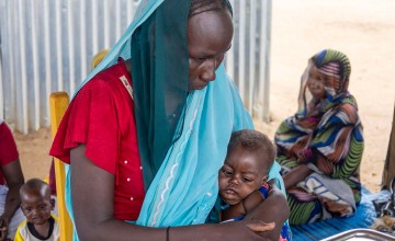 A woman wearing a blue headscarf and red dress sits holding a young child in her arms at a health or nutrition centre. Other women and children sit nearby in the background, with basic medical supplies visible on a table.