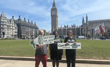 Three Concern staff stand in front of the Houses of Parliament holding signs saying Defend UK Aid.