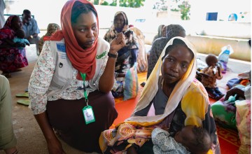 A mother sit on the ground with a baby on her lap, she is talking to a Concern healthcare worker and there are other mothers with their babies around them. 