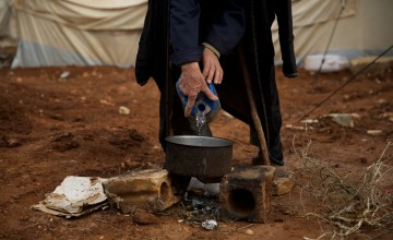 A man pours water from a jug into a pot that is held up by two small bricks over a burnt pile of sticks.