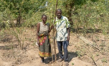 Two people stand together in a dry, sparsely vegetated field, surrounded by small trees and plants. One is wearing a patterned skirt and sleeveless top, and the other is dressed in a light shirt and dark trousers. They hold or stand beside a tall, slender plant, with sunlight casting shadows across the ground.