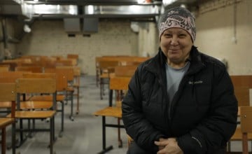 A person wearing a winter jacket and patterned hat sits in a room filled with rows of wooden school chairs. The setting appears to be a basement shelter with exposed brick walls and overhead ventilation ducts.