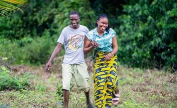  Two people walk, smiling together through a grassy, open area bordered by dense green vegetation. One person wears a grey T‑shirt and light shorts, while the other wears a patterned yellow and blue skirt and a teal top. They hold onto each other’s arms as they move forward across the uneven ground.