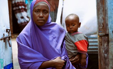 A woman wearing a purple wrap sits holding a young child on their lap. The child is dressed in a grey and red top. They are seated outdoors between structures made of corrugated metal and wooden materials. The scene appears to be in a modest residential or settlement area with various fabrics and materials visible in the background.