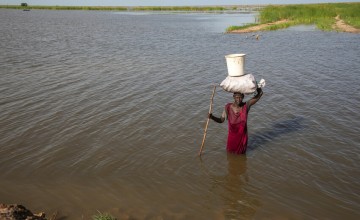 A person stands waist‑deep in a wide body of water, using a long wooden stick for support. The person balances a large white container placed on top of a wrapped bundle carried on their head. The water stretches far into the distance, bordered by green vegetation along the shoreline under a bright sky with scattered clouds.
