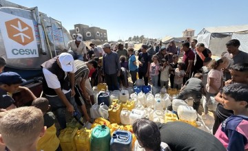 A large group of people gathers around a water truck as workers fill numerous plastic jerry cans with water in a camp-like setting under bright sunlight.