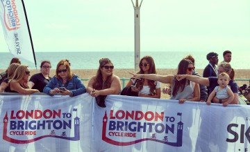 A group of spectators stand behind a barrier in Brighton, pointing and watching participants, with London to Brighton Cycle Ride signage visible.