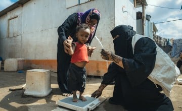 A child stands on scales while being held by his mother and a woman health worker examines his weight
