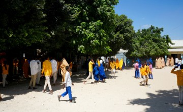 Students in colorful uniforms walk and gather under large trees in a sunny school courtyard.