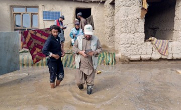 Two men walk through flooding at shin-level in front of a home