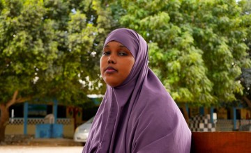 Young woman in a purple headscarf sitting outdoors with trees and buildings in the background.