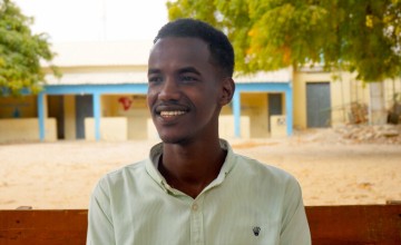 Smiling young man in a light shirt sitting on a bench outdoors with trees and buildings behind.