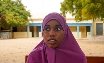 Girl in a purple headscarf sitting on a bench outdoors with trees and buildings behind.