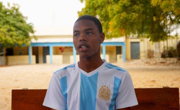 Young man in a white and light blue jersey sitting on a bench outdoors with trees and buildings behind.
