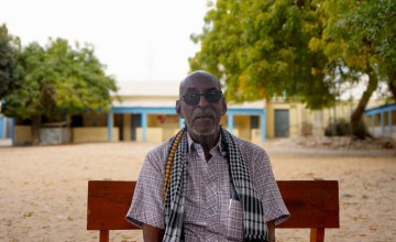 Older person wearing sunglasses sits on a bench outdoors with trees and buildings in the background.