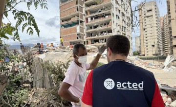 Aid worker speaks with a resident beside rubble and a damaged apartment building after a disaster.