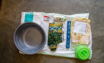 A newborn care kit is neatly arranged on a mat, including a baby blanket, clothing, soap, baby lotion, a basin, a covered bucket, fabric wraps and other essential hygiene items provided to support mothers and newborns.