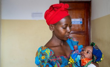 A woman wearing a bright red headscarf and colourful patterned dress cradles her baby in her arms while looking down with care. Wrapped in a vibrant blanket, the baby looks back up at her inside a healthcare facility.